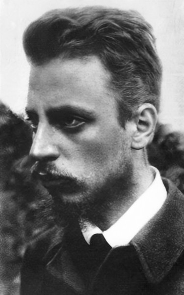 Portrait of the poet Rainer Maria Rilke in 1900, a young man with swept back hair and a light moustache, in a dark coat and white collar, looking to the side.