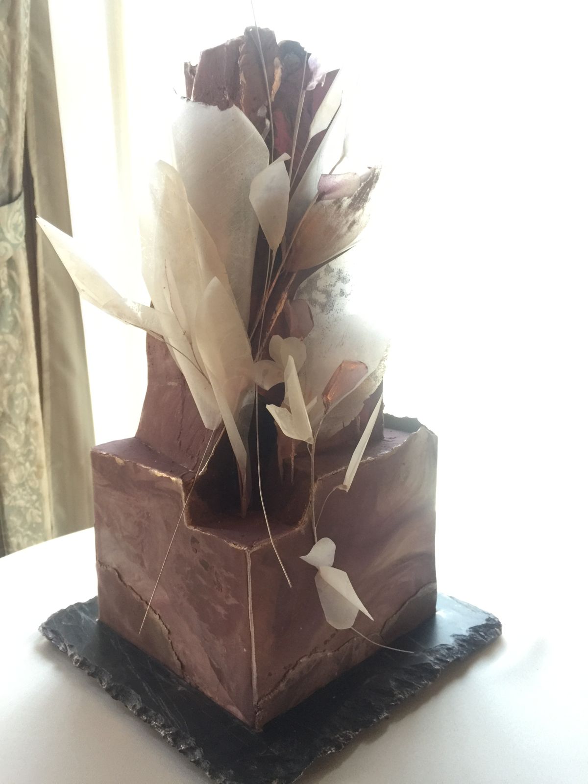 The finished cake seen from a low angle in window light: a plum and copper marbled block with a tall jagged fin rising from it, hung with pale wafer paper petals.