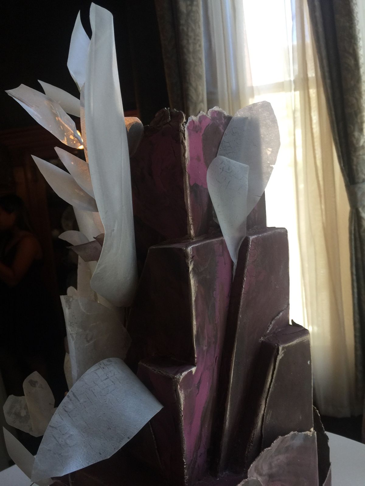 A close view of the top of the cake: layered plum blocks with a pink vein, and curled sheets of wafer paper in grey and white beside a curtained window.