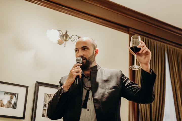 A bald man in a dark jacket raising a glass of red wine as he speaks into a microphone.