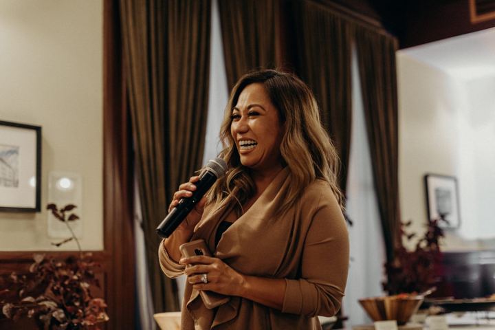 A woman with long highlighted hair in a brown wrap top, laughing as she speaks into a microphone.