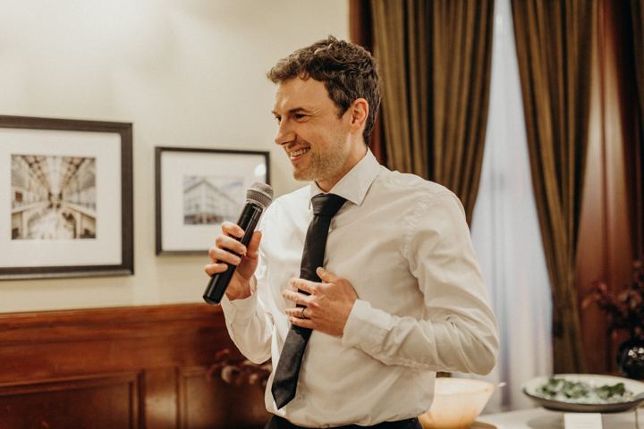 John Brian Kirby in a white shirt and dark tie, laughing as he speaks into a microphone, one hand held out.