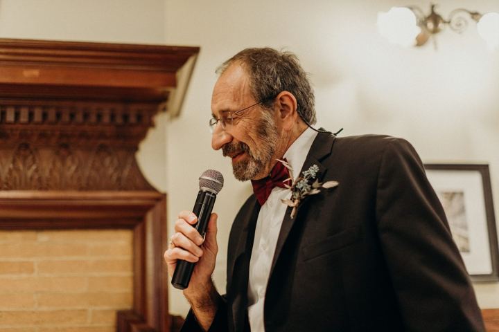 A man in a dark suit with a red bow tie and a boutonniere, smiling as he speaks into a microphone.