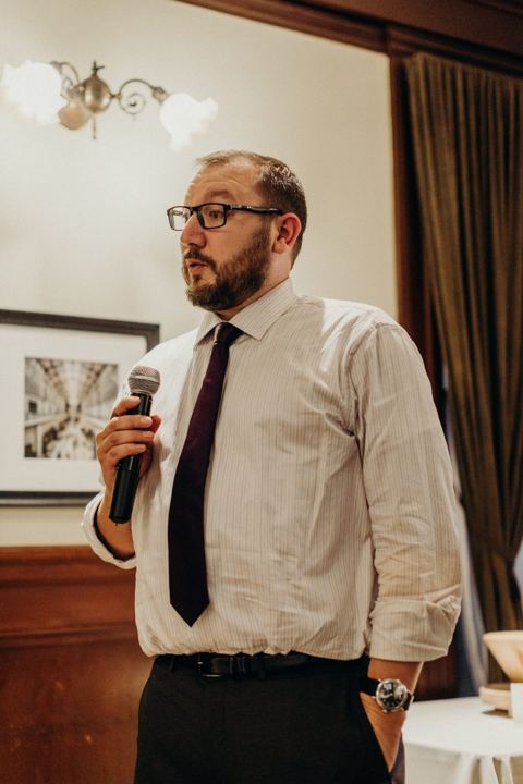 A man in glasses and a striped shirt with a dark tie, standing with a microphone in the dining room.
