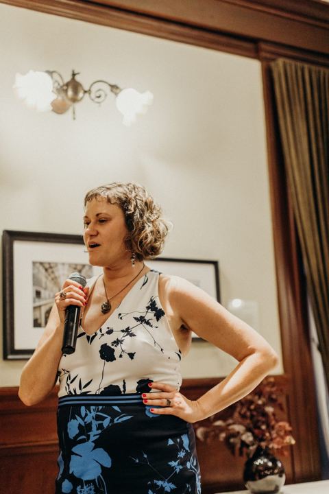 A woman with short curly hair in a blue and white patterned dress, one hand on her hip, speaking into a microphone.