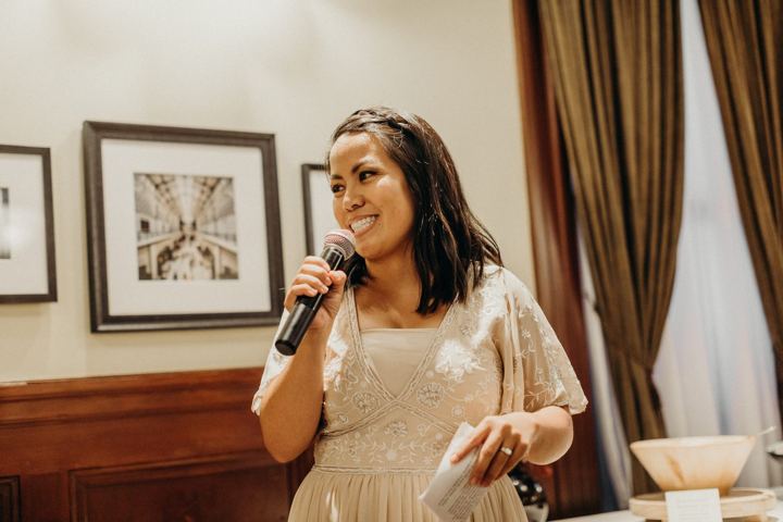 A woman with dark hair in a pale beaded dress, smiling as she speaks into a microphone.