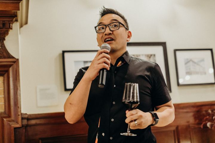 A man in glasses and a black shirt speaking into a microphone, a glass of red wine in his other hand.